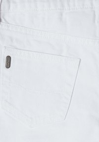 Vit denimtyg har en bakficka med ett subtilt texturerat mönster och en liten, silverfärgad märkesetikett sydd i kanten.