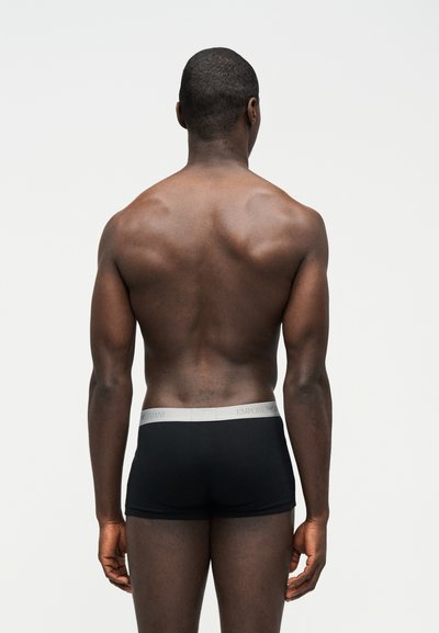 Lihaseline mees seisab seljaga, kandes musti bokserpükse heleda halli vöökohaga, millel on brändinimi "Emporio Armani", taust on lihtne.