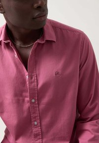 Camisa de botones de algodón en rosa oscuro, con un logo en el pecho, cuello clásico y botones en azul claro. Textura suave y ajuste entallado.