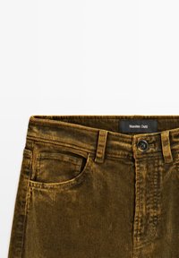 Pantalons en velours côtelé marron avec une texture lisse, des côtes visibles, un design à cinq poches, une fermeture à bouton et une étiquette noire portant l'inscription "Massimo Dutti."