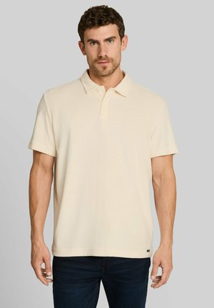 Man with short curly hair wearing a cream-colored short-sleeve polo shirt and dark pants against a plain gray background.