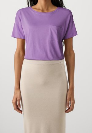 Mujer que lleva una camiseta de manga corta color lavanda con un bolsillo en el pecho, metida dentro de una falda de cintura alta beige, sobre un fondo liso.