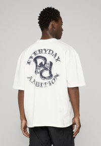 Vit t-shirt med korta ärmar och en rund grafisk slogan "EVERYDAY AMBITION" i marinblått, med ett slitet tryck och avslappnad passform.