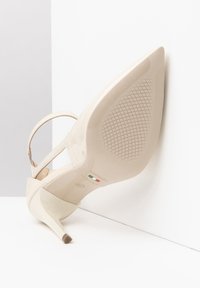 Scarpa con tacco alto in pelle verniciata beige, con punta affusolata, sottile cinturino alla caviglia e suola testurizzata con motivo antiscivolo.