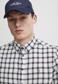 Svart og hvit rutete skjorte med krage og knappestolpe, med mørke knapper og en liten logo på brystet. Brukt med en marineblå caps.