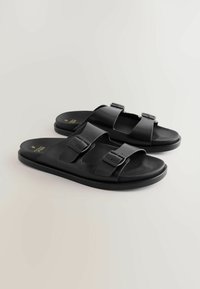 Sandales slides noires avec deux brides réglables à boucle et une semelle plate, placées côte à côte sur un fond clair.