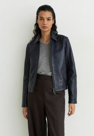 Frau mit dunklem lockigem Haar, die eine schwarze Lederjacke, ein graues Shirt und dunkelbraune, hoch taillierte Hose vor einer weißen Wand trägt.