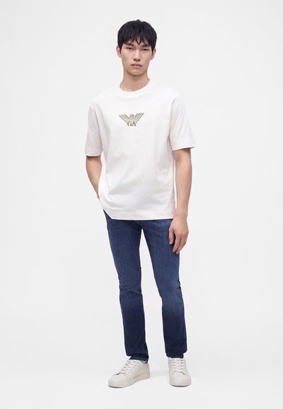 Νέος άνδρας που φοράει λευκό κοντομάνικο T-shirt με λογότυπο, μπλε τζιν και λευκά αθλητικά παπούτσια, στέκεται με το ένα χέρι στην τσέπη σε απλό φόντο.