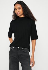 Top negro de punto acanalado con mangas cortas y cuello alto, combinado con jeans gris oscuro. Textura suave, diseño ceñido al cuerpo, acentos mínimos.