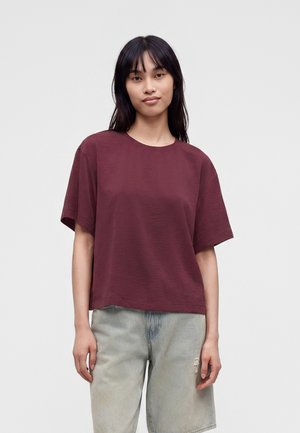 Ung kvinde med glat sort hår iført en løs bordeaux kortærmet skjorte og lysegrå denimshorts står foran en ensfarvet baggrund.