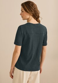 Dunkelblaues T-Shirt mit kurzen Ärmeln und rundem Ausschnitt, glatter Textur und einem dezenten Nahtdetail am Rücken, kombiniert mit hellen Hosen.