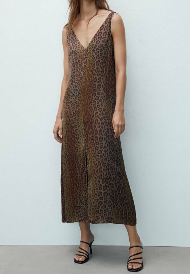 Femme portant une robe midi sans manches à col en V, imprimé léopard, avec des sandales noires à talons hauts et brides, debout contre un mur blanc uni.