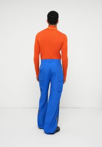 Camisa polo laranja de mangas compridas com um colarinho estreito. Calças cargo azuis largas com bolsos laterais e riscas amarelas em contraste ao longo das costuras.