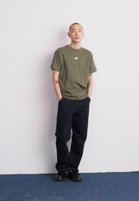 Olive zaļš Nike T-krekls ar baltu logotipu, apvienots ar melniem džinsiem un melniem apaviem, stāvot pretī vienkāršai baltai fona.