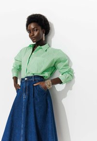 Chemise boutonnée rayée verte et blanche à manches longues, associée à une jupe maxi en denim bleu foncé. Accessoirisée avec un bracelet à plusieurs brins.
