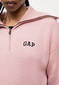 Personne portant un pull en maille côtelée rose avec un col à demi-fermeture éclair et un logo "GAP" noir brodé sur la poitrine.
