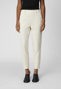 Pantalon ajusté de couleur crème avec coupe slim, plis à l'avant, longueur raccourcie et tissu texturé. Porté avec des bottines noires.