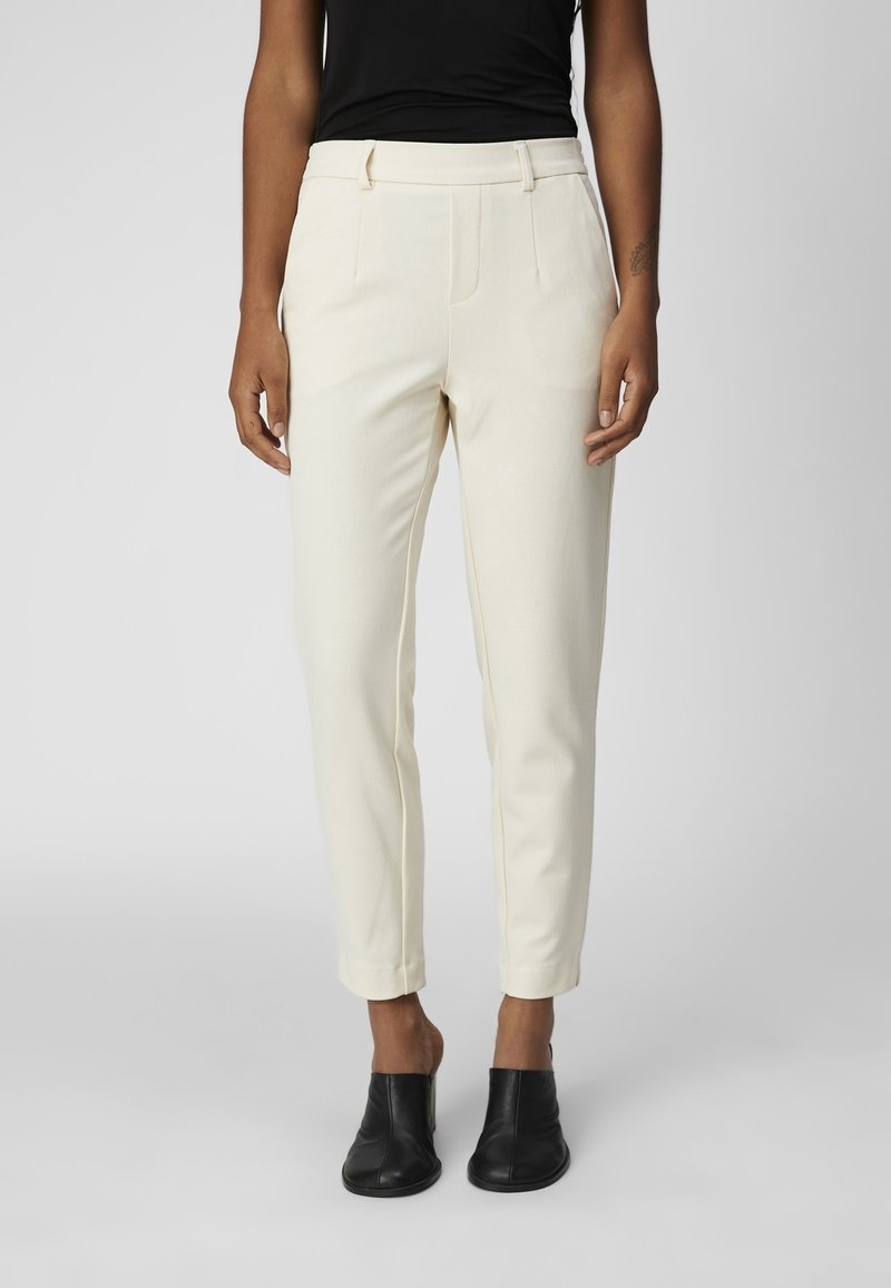 Pantalon ajusté de couleur crème avec coupe slim, plis à l'avant, longueur raccourcie et tissu texturé. Porté avec des bottines noires.