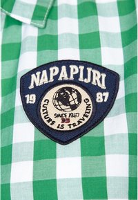 Grünes und weißes kariertes Gewebe mit einem gestickten marineblauen Abzeichen, das einen Globus, den Text "Napapijri" und "Seit 1987" sowie Designelemente zeigt.