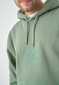 Green hoodie made of soft fabric featuring a front pocket and drawstrings, with a light blue embroidered logo on the chest.