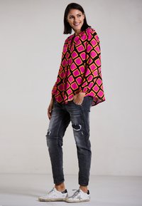 Farbige Langarmbluse mit einem pinken und gelben Rautenmuster sowie schwarzen Akzenten, kombiniert mit zerrissenen schwarzen Jeans und weißen Sneakers.