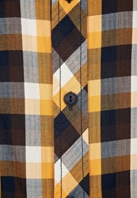 Närbild av randigt tyg i gult, brunt, svart och vitt med svart kn button och synlig söm på skjortans framplacket.