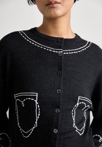 Fekete kötött kardigán, kerek nyakkal, elülső gombos záródással, fehér varrással a zsebek és a gallér körül.