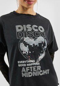 Czarny t-shirt z grafiką serca w kształcie kuli dyskotekowej w kolorze srebrnym, z białym nadrukiem "DISCO DISCO" oraz "WSZYSTKO DOBRE DZIEJE SIĘ PO PÓŁNOCY."