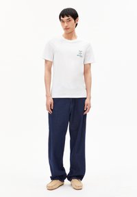 Weißes Baumwoll-T-Shirt mit kurzen Ärmeln und einem dunkelgrünen Grafikdesign, kombiniert mit weit geschnittenen dunkelblauen Jeans und beigen Schuhen.