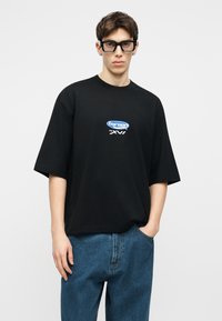 Jeune homme portant un t-shirt noir oversize avec un petit logo blanc et bleu, un jean bleu et de grandes lunettes carrées noires, debout avec une main dans la poche.
