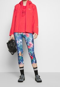 Klar rød vandtæt jakke, tætsiddende blomster leggings med blå baggrund og sort cykelhjelm; har også mørke sko og sokker.
