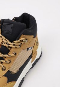 Bota de senderismo alta con parte superior sintética color marrón claro, detalles en negro, paneles de malla y cordones beige. Presenta una suela negra robusta para mayor tracción.