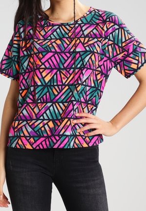 Frau trägt ein kurzärmeliges Oberteil mit buntem, geometrischem Muster und schwarze, hoch taillierte Jeans, Hand auf der Hüfte ruhend.