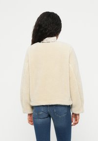 Lys cremefarvet faux fur jakke med krave, drop-skuldrer og afslappet pasform, parret med blå jeans. Blød tekstur og ingen synlige lukninger.