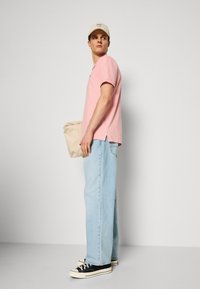 Junger Mann, der ein pinkes T-Shirt, hellblaue Jeans, schwarze Sneakers und eine beige Kappe trägt und eine beige gefaltete Tasche vor einem einfarbigen Hintergrund hält.