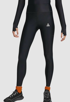 Leggings de sport noirs avec une texture lisse, taille haute et design ajusté ; présentent un petit logo blanc sur la cuisse gauche.