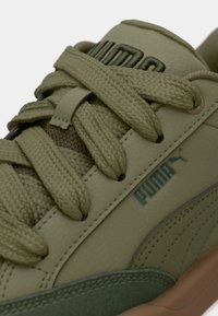 Tuvinājums ar olīvu zaļajiem Puma snickers ar bieziem šņorēm, izcilotu logo uz mēles un strukturētu zoli brūnos un zaļos toņos.