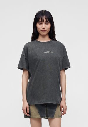 Jeune femme aux longs cheveux foncés porte un T-shirt ample gris foncé et une courte jupe en jean bicolore, debout devant un fond clair uni.