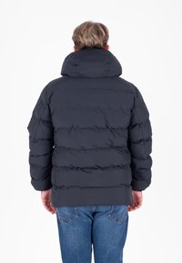 Donkergrijze pufferjack met een hoge kraag en horizontale stiksels, gladde textuur en elastische manchetten. Draagt over blauwe jeans.