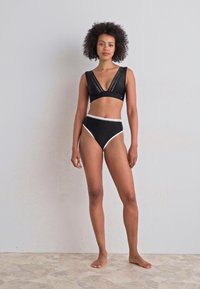 Svart bikinitopp med teksturert svart del med utskjæringer og dyp V-hals. Høyt midje-bunn med hvit kant. Glatt stoff, minimalistisk design.