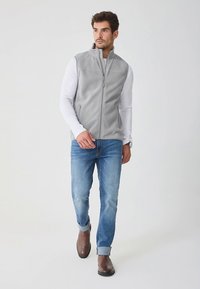 Vestă gri de fleece cu guler înalt și fermoar frontal, purtată peste o cămașă albă cu mâneci lungi, asortată cu blugi albaștri și cizme maro până la gleznă.