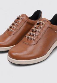 Zapatos de cuero marrón con cordones elásticos, puntera redondeada y suela de goma. Presentan una entresuela de color crema en contraste y detalles de costura sutiles.
