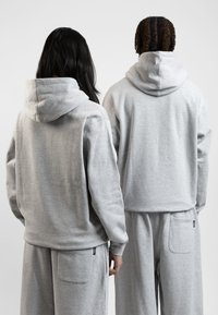 Světle šedé mikiny vyrobené z měkkého materiálu, s klokaními kapsami a volným střihem, zobrazené ze zadní strany s odpovídajícími kalhotami.