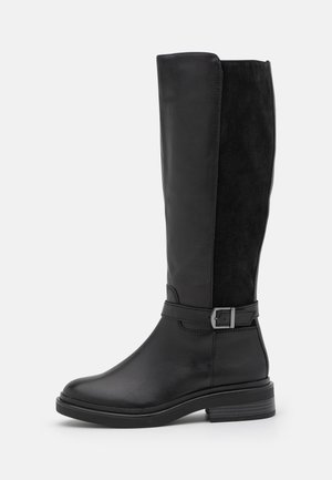 Botte noire montante jusqu'au genou avec devant en cuir, dos en daim, petit talon, bout rond, et boucle décorative sur le côté sur fond gris.