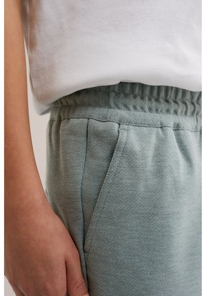 Prim-plan cu o persoană purtând pantaloni cu talie elastică de culoare albastru deschis și o cămașă albă băgată în pantaloni, cu o mână relaxată pe lângă șold.