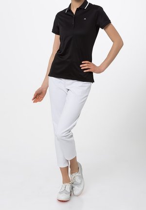 Czarna koszulka polo z kołnierzykiem, białe akcenty; zestawiona z białymi, krótkimi spodniami i jasnoszarymi butami. Prosty design, gładka faktura.
