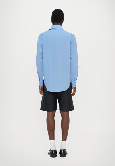 Chemise à manches longues bleu clair avec un ourlet incurvé et des poignets boutonnés, portée avec un short noir et associée à des chaussures noires et des chaussettes blanches.