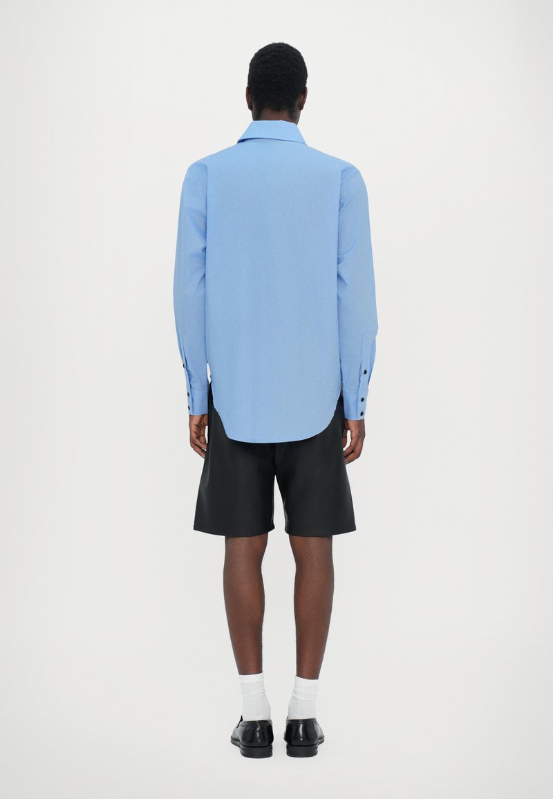 Lyseblå langærmet skjorte med buet kant og knapærmer, båret med sorte shorts og parret med sorte sko og hvide sokker.