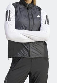 Czarna i biała kurtka sportowa z zamkiem na przodzie, wysokim kołnierzem i trzema czarnymi paskami na każdym rękawie. Wykonana z lekkiego, teksturowanego materiału.