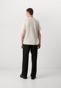 Lys grå t-skjorte med korte ermer laget av bomull, kombinert med svart vide bukser. Modellen står med en tatovering synlig på armen.